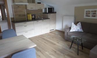 A modern kitchen with wood furniture and a cozy living room. There is a table, chairs, and a sofa with a decorative cushion.