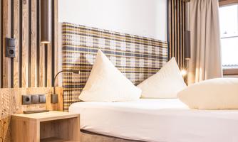 A cozy hotel room with a large bed and modern wooden decor. The walls are decorated with wooden panels and there are natural light sources through the windows.