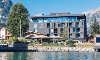 A modern hotel by the water with large windows and sun umbrellas. In the background, impressive mountains can be seen.