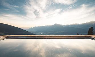 A quiet pool with a wooden terrace and a stunning view of the mountains. The clouds reflect in the water, creating a peaceful atmosphere.