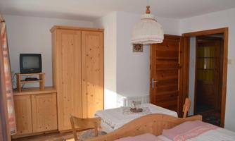 A simple bedroom with two beds and a wooden table. The walls are bright, and there is a wooden wardrobe and a lamp.