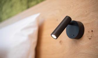 A modern wall light made of black metal. It is mounted on a bright wooden wall above a bed.