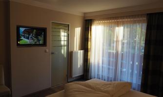 A bright hotel room with a television on the wall and large windows. The curtains are open, allowing plenty of daylight in.
