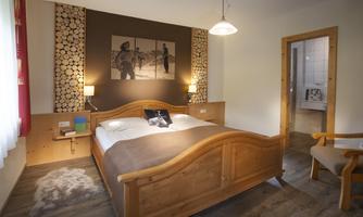 A cozy bedroom with a large wooden double bed and warm colors. Decorative pictures hang on the wall, and there is a chair in the corner.