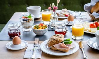 A beautifully set breakfast table with fresh rolls, cold cuts, yogurt, fruit, and various drinks. The table also has eggs and jam in small jars.