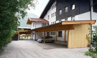 A modern building with wooden cladding and a covered parking lot. In the foreground, several cars are parked on gravel.