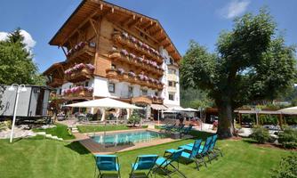 A beautiful hotel with wooden cladding and blooming balconies. In the foreground, there is a pool surrounded by sun loungers and a green meadow.