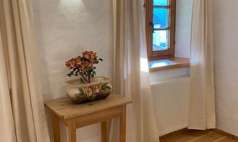 A bright room with wooden flooring and a small table. Next to the window are curtains and a flower vase.