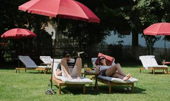 Two people are relaxing on loungers under red sun umbrellas. The surroundings are green and inviting, ideal for relaxing.