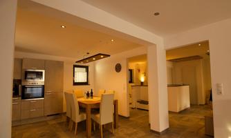 A modern dining room with a wooden chair and an elegant table. The kitchen is well-equipped and warmly lit.
