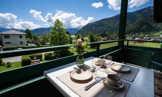 A beautiful table on a balcony with a view of the mountains. The table decoration includes a flower vase, glasses, and dishware.