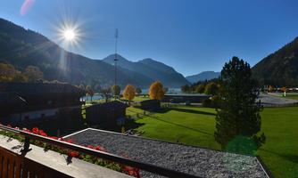 A picturesque mountain landscape with a bright sun and clear blue sky. In the foreground, a green meadow and some trees.
