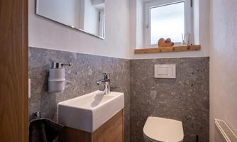 A modern bathroom with gray stone walls and a wooden element. It features a sink, a toilet, and a window with natural light.