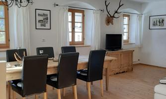 A cozy dining area with a long table and black chairs. Large windows let in plenty of light, and there is a wooden ceiling with a stylish chandelier.