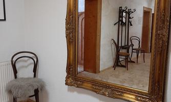 A large, golden mirror hangs on the wall and reflects a hallway-like room. Next to the mirror stands a chair with a fur cushion and a rug with the inscription "Wonderful to have you here."