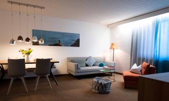 A modern living room with a cozy sofa, a dining table, and stylish lamps. Large windows let in light and create an inviting atmosphere.