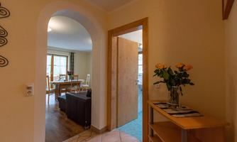 An inviting entrance area with an open door to a bright room. Fresh flowers are on a shelf, and there is a cozy seating area.