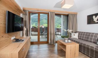 A modern, bright living room setup with a sofa, a wooden cabinet, and a television. Large windows offer a view of the surrounding nature.