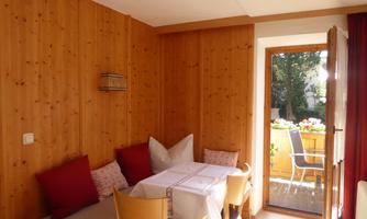 A cozy room with wooden walls, a table, and several chairs. Through the open door, you can see a lovely outdoor area with garden furniture.