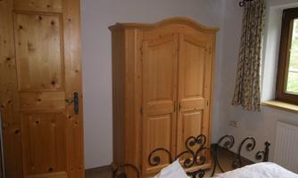 A cozy bedroom with a wooden wardrobe and a metal bed. The walls are light and a window with curtains provides daylight.