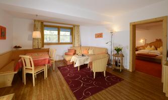 A bright and inviting living room with a dining table and comfortable armchairs. In the background, a bedroom is visible, furnished with a double bed.