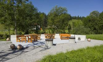 A cozy outdoor area with wooden furniture and a concrete floor. Surrounded by trees and a green meadow, perfect for relaxation and social gatherings.