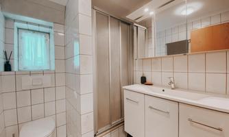 A modern bathroom with white tiles and a shower. There is a sink and a toilet as well as a small window with a view.