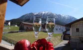 Two glasses of sparkling wine are on a table, surrounded by a red apple and a rose. In the background, snow-covered mountains and a tranquil landscape can be seen.