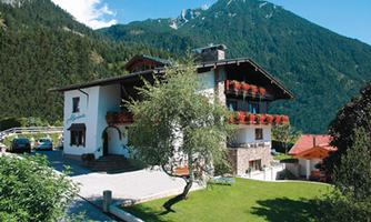 A charming house in the mountains with balconies and a well-maintained garden. In the background, high mountains and a clear blue sky can be seen.