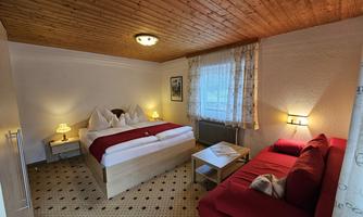 A cozy bedroom with a double bed and a red sofa. The room has a wooden ceiling and a window with curtains.