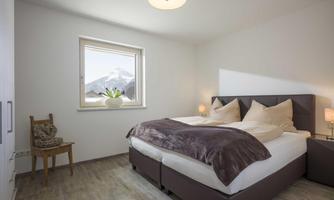 A bright bedroom with a large bed and cozy bedding. A pot with plants sits by the window and you can see the mountains through the window.