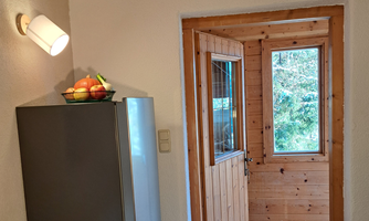 A kitchen with a refrigerator and a fruit bowl on the countertop. In the background, there is a wooden-paneled door and a window with a view of the outdoors.