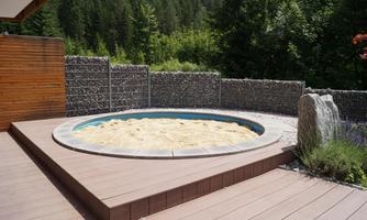 A round sand area surrounded by a wooden deck and natural stone walls. In the background, trees and a green landscape can be seen.