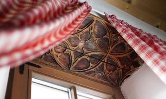 A window with an artistically carved wooden ceiling and red-and-white checked curtains. The atmosphere is cozy and rustic.