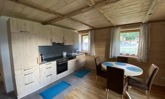 A modern kitchen in a wooden house with a dining table and chairs. Large windows let in plenty of light and offer a view outside.