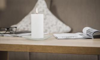 An elegant candle stands on a table with a glass base. Next to the candle are a pair of glasses and some magazines.