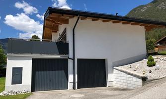 A modern house with a garage area and a terrace. It is located in a rural setting with mountains in the background.