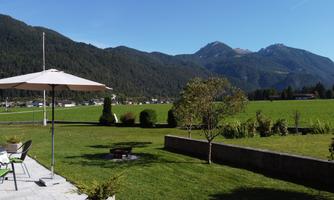 A beautiful landscape with green meadows and mountains in the background. In the foreground, there is a pavilion and a fire pit.