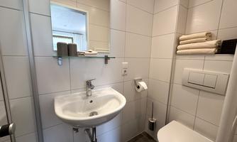 A small, modern bathroom with white tiles and a sink. Towels are neatly stacked on a shelf, and a window provides daylight.