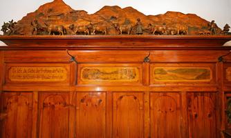 A decorative wooden panel with carved figures of people and animals. In the background, stylized mountains are depicted.