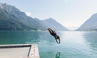 A man jumps from a dock into a clear, blue lake. In the background, majestic mountains and a bright blue sky can be seen.