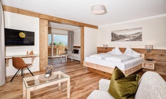 A modern hotel room with wooden elements and large windows. It features a double bed, a desk, and a cozy seating area.
