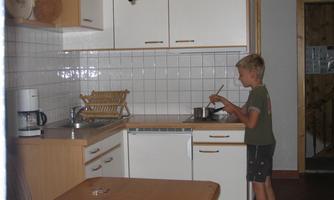 A boy stands in a modern kitchen and stirs in a pot. The kitchen is bright and tidy, with wooden shelves and white cabinets.