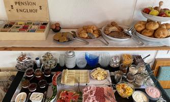 A rich breakfast buffet with various types of bread, cold cuts, jam, and fresh fruit. There are also yogurt and multiple types of cheese available on the table.