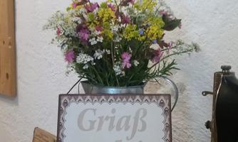 A beautiful bouquet of flowers in a metal can is on a table. Next to it lies a sign with the inscription "Griaß enk!".
