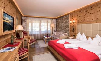 A cozy room in chalet style with wooden paneling and warm colors. There is a double bed, a living area, and plenty of daylight through large windows.