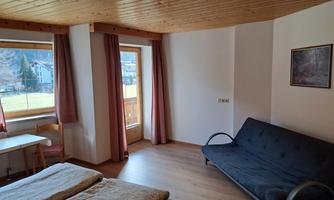A simple room with wooden paneling, a double bed, and a sofa bed. Large windows provide plenty of daylight and a view of the outside.