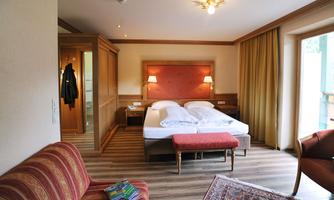 A cozy hotel room with two single beds and a red sofa. The furniture is made of wood, and there are large windows with curtains.