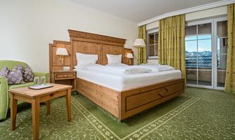 A cozy hotel room with a large bed, a table, and an armchair. Large windows offer a view outside and the curtains are in a fresh green.
