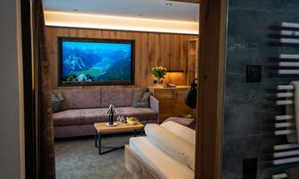 A cozy hotel room with a sofa, a table, and a large television. The walls are made of wood and there are fresh flowers on the table.